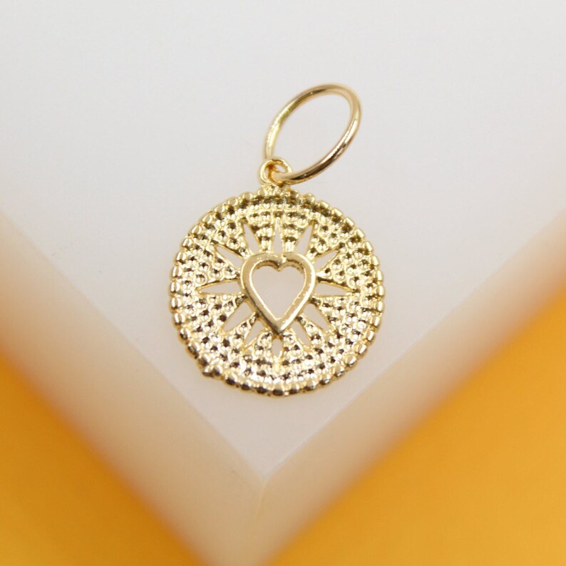 A delicate gold pendant featuring an openwork heart design inside a textured circular frame rests on a white display surface.