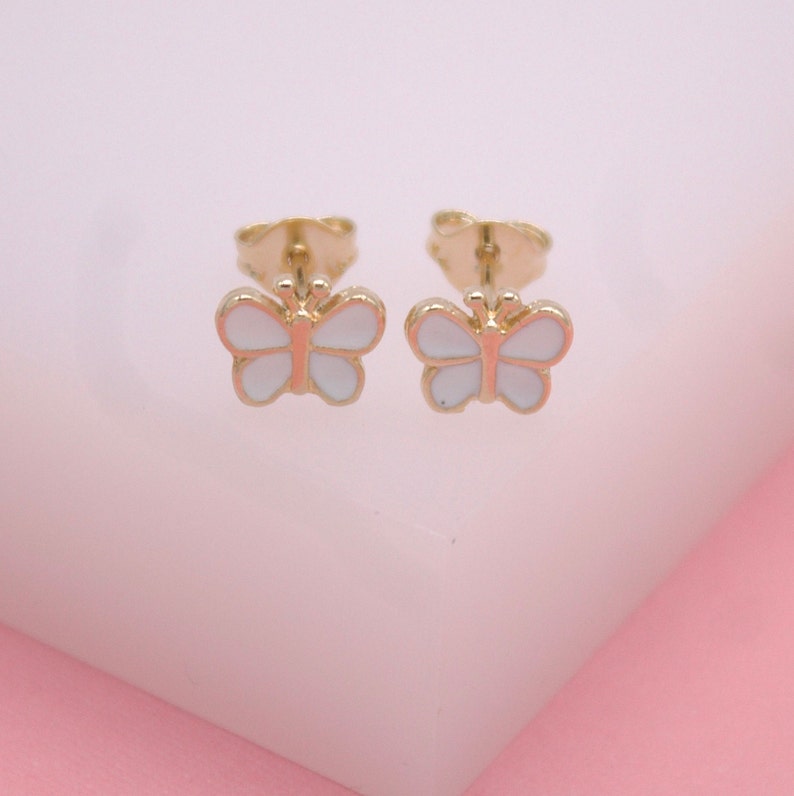 A pair of gold butterfly stud earrings features delicate white enamel wings displayed against a soft pink background.