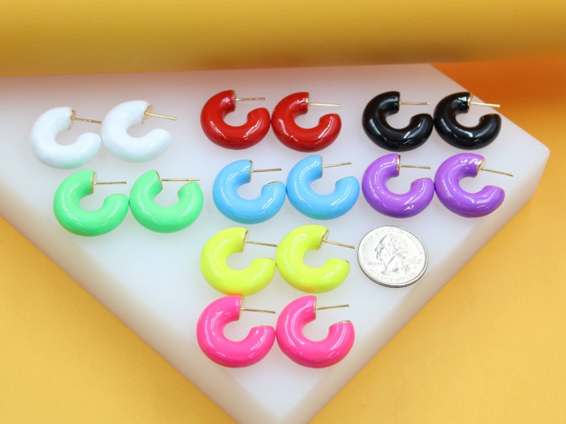 An assortment of colorful enamel huggie hoop earrings displays pairs in white, red, black, green, blue, purple, yellow, and pink, arranged on a white display stand next to a US quarter coin for size comparison.