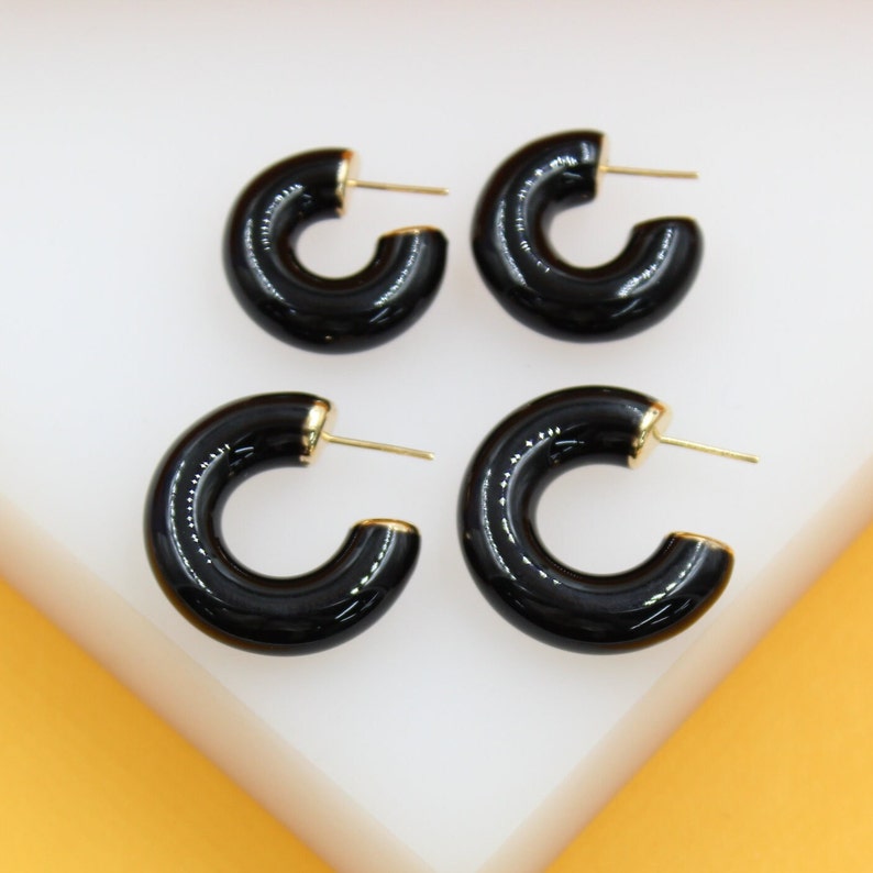 Two pairs of glossy black enamel hoop earrings with gold-plated posts rest on a white surface.