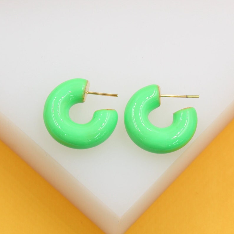 A pair of vibrant neon green enamel hoop earrings rests on a white and yellow display stand.