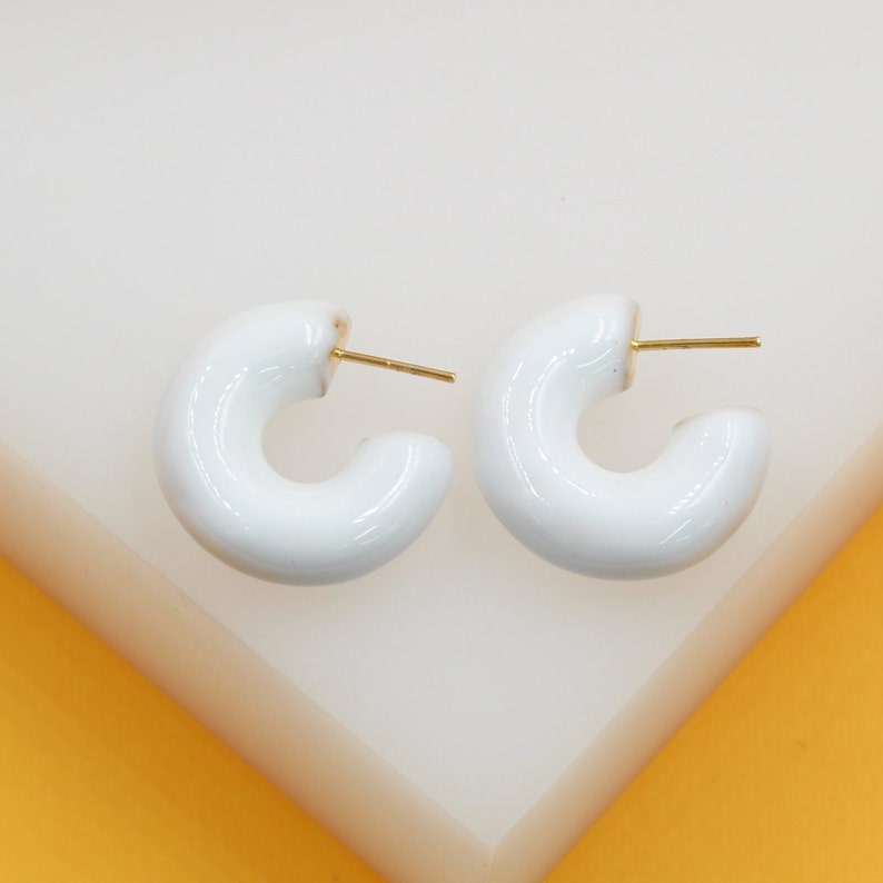 A pair of glossy white thick hoop earrings with gold posts rests on a white display surface.