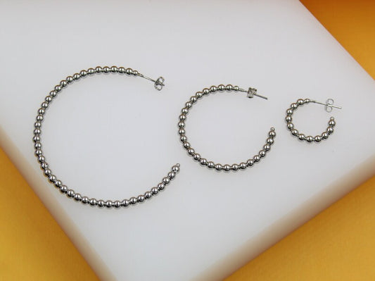 Three graduating sizes of silver ball-beaded hoop earrings rest on a white surface against an orange background.