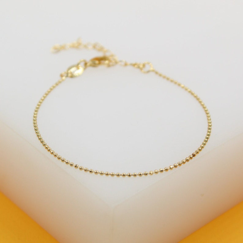 A delicate gold bead chain bracelet rests on a clean white display surface against a warm yellow background.