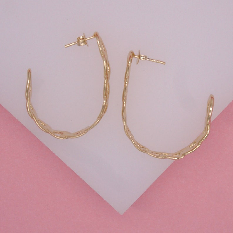 A pair of textured gold hoop earrings rests on a pink and white surface.