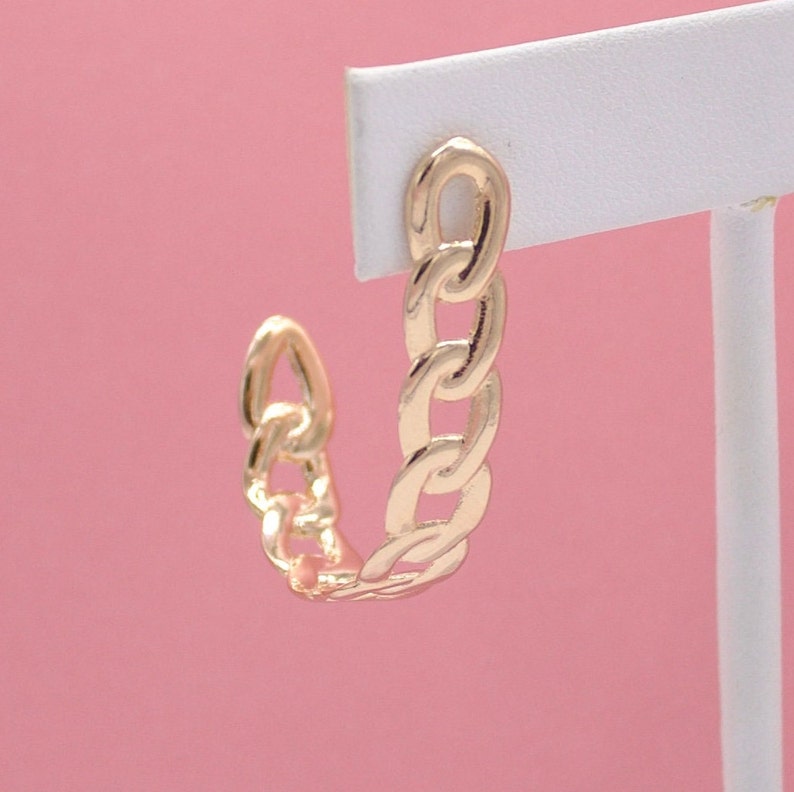 A gold chain-link hoop earring hangs on a white display card against a pink background.
