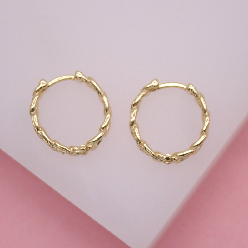 Two gold twisted hoop earrings rest on a pale pink display block.