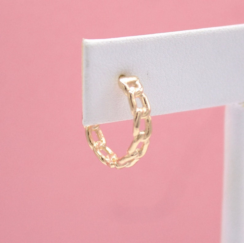A gold chain-link hoop earring is displayed on a white card against a pink background.