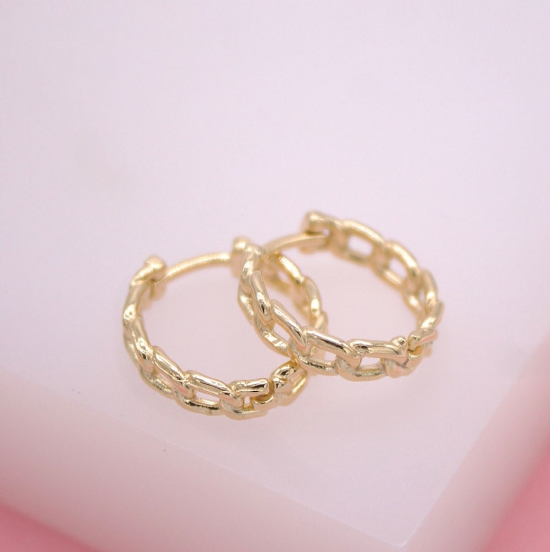 A pair of gold chain link huggie hoop earrings rests delicately on a soft pink surface.