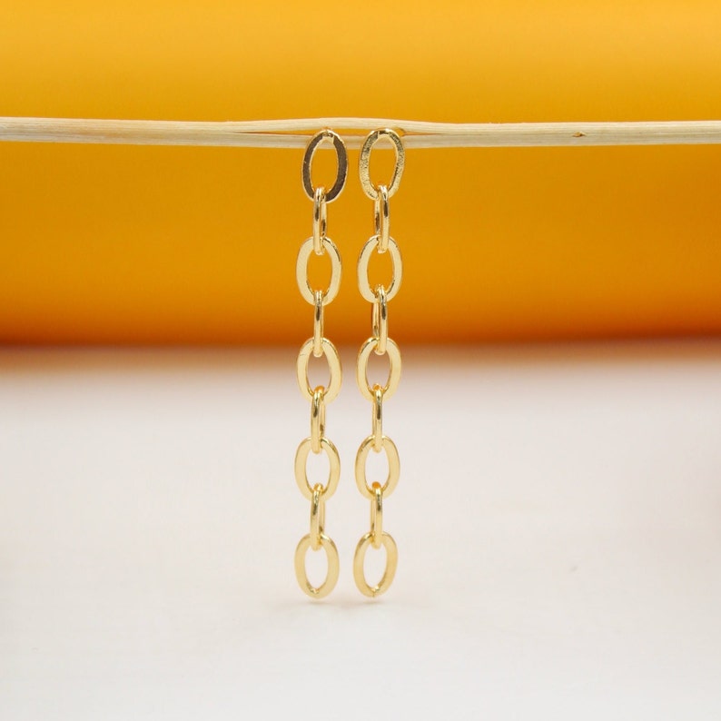 Two long, gold-plated chain drop earrings hang gracefully from a horizontal wooden stick against a bright yellow and white background.