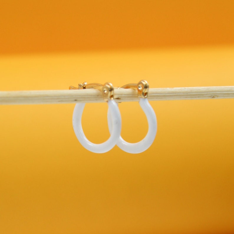 A pair of gold hoop earrings with white enamel coating hangs delicately from a wooden stick against a warm yellow background.
