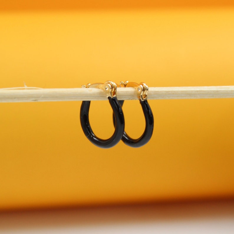 A pair of small gold hoop earrings with black enamel coating hangs delicately from a wooden stick against a warm yellow background.