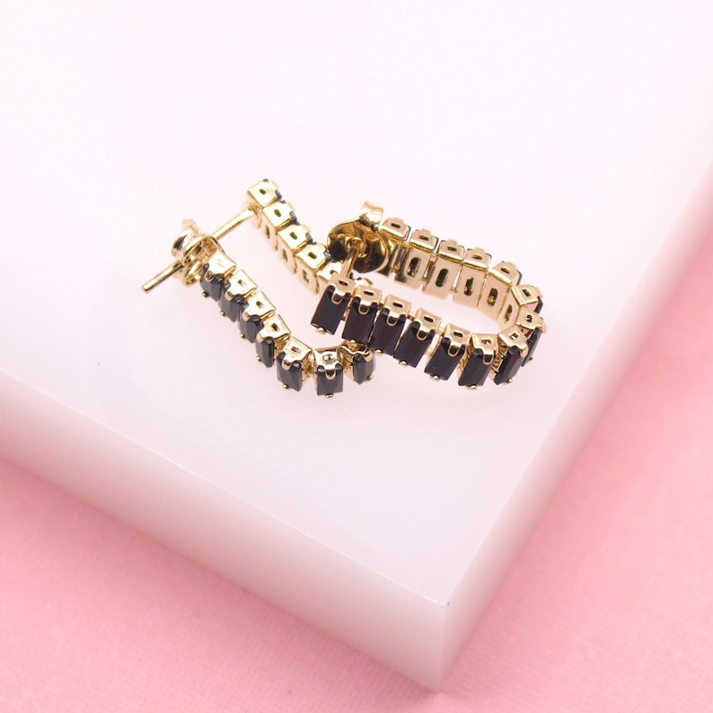 A pair of gold huggie hoop earrings featuring dark rectangular stones rests on a white and pink surface.