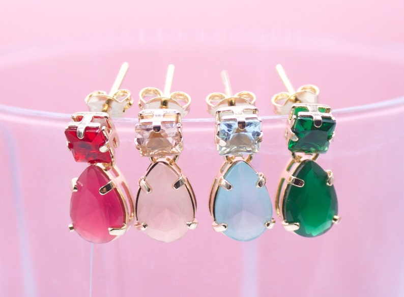 Four pairs of colorful gemstone drop earrings in red, light pink, light blue, and green rest against a soft pink background.
