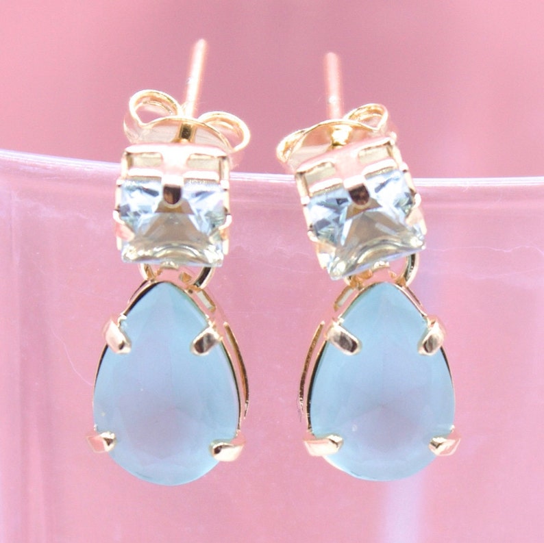 A pair of elegant gold-plated drop earrings features square blue stones at the top and dangling pear-shaped blue gems against a soft pink background.