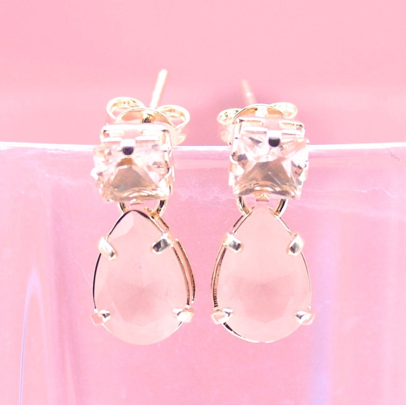 A pair of delicate gold earrings features square top crystals suspended above teardrop-shaped pink gemstones against a soft pastel background.