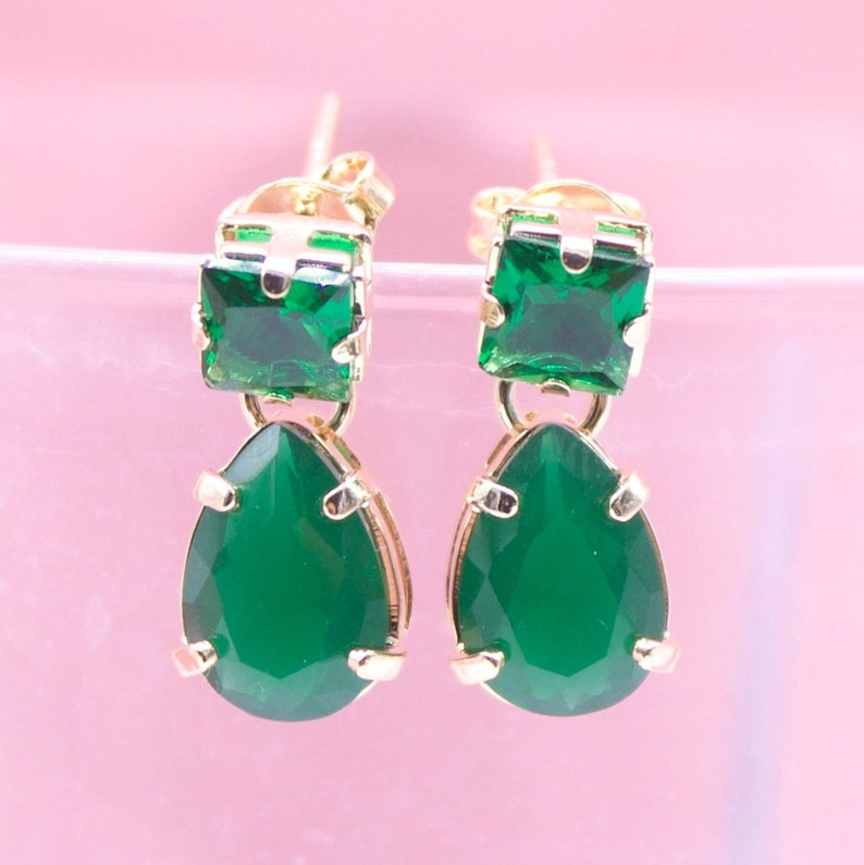 A pair of gold earrings featuring emerald-cut green crystal studs and dangling teardrop-shaped green stones rests against a soft pink background.