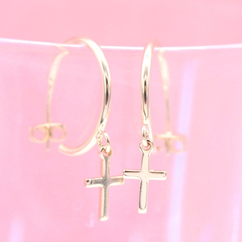 A pair of gold hoop earrings with dangling cross charms rests against a soft pink background.