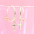 A pair of gold hoop earrings with dangling cross charms rests against a soft pink background.