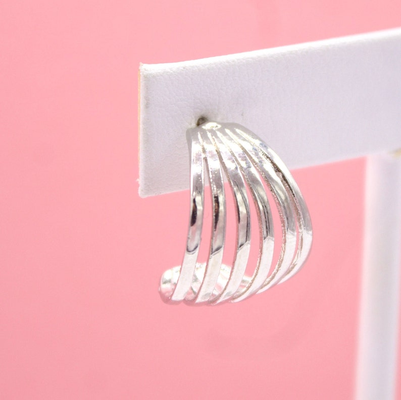 A polished silver multi-band huggie hoop earring is displayed on a white jewelry card against a soft pink background.