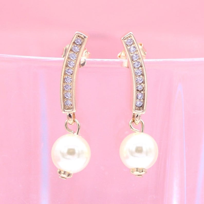 A pair of gold earrings featuring curved bars encrusted with sparkling crystals and dangling white pearl drops rests against a soft pink backdrop.