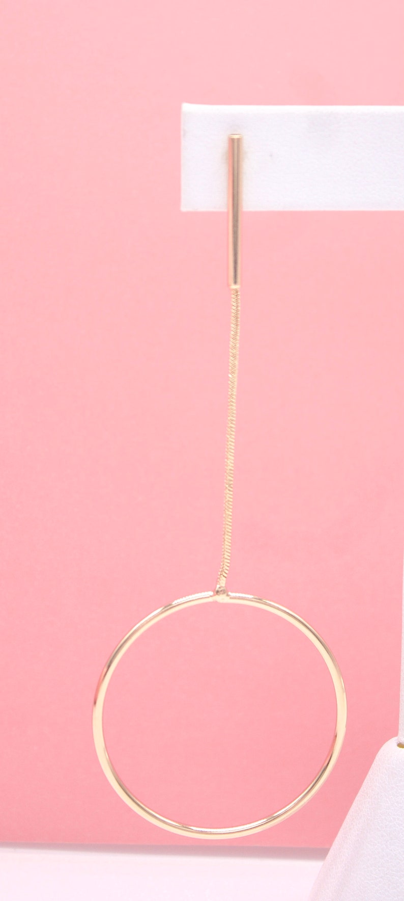 A modern gold drop earring features a sleek bar connected to a delicate chain and a circular hoop against a soft pink background.
