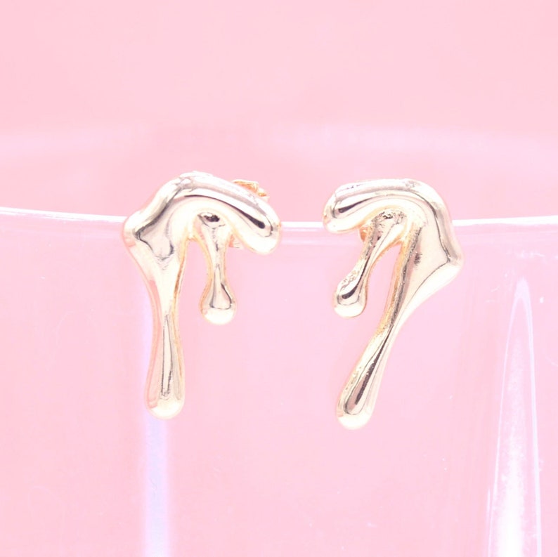 A pair of gold drip-design stud earrings rests delicately against a soft pink background.