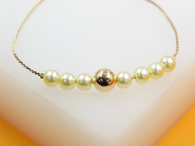 A delicate gold chain bracelet features alternating lustrous pearls and a polished central gold bead resting on a white surface.