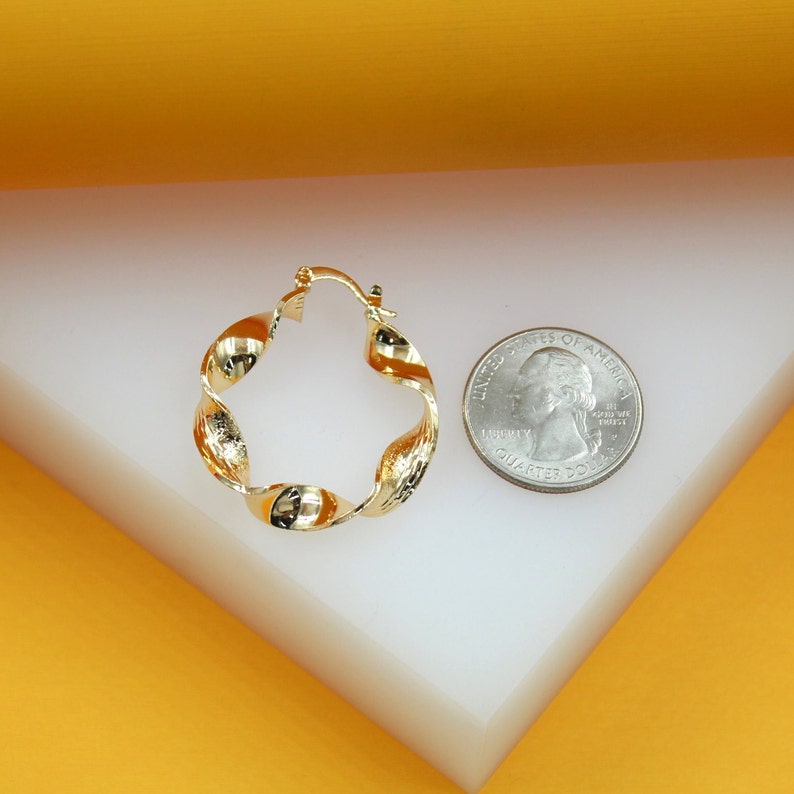 A polished gold twist hoop earring rests next to a US quarter on a white display surface.