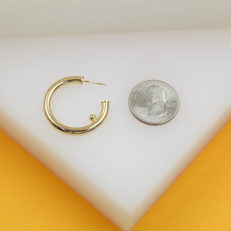 A gold hoop earring rests beside a US quarter for size comparison on a white display surface.