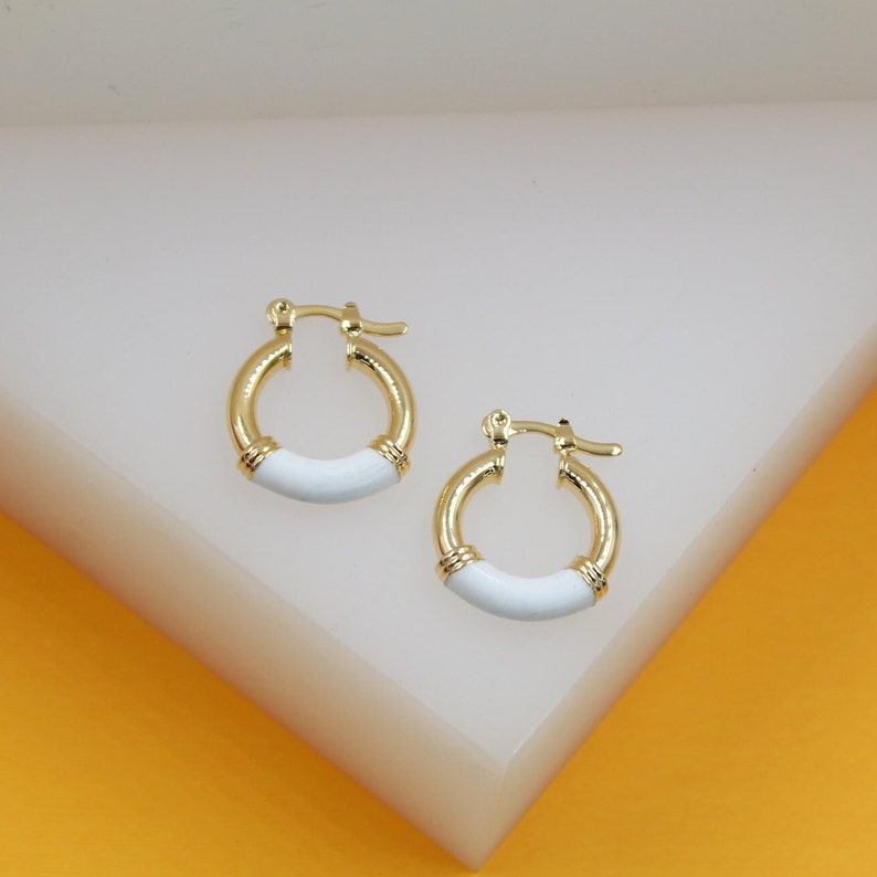 A pair of gold huggie hoop earrings featuring white enamel accents rests on a clean white display surface.
