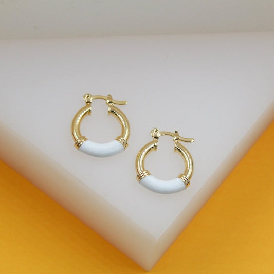 A pair of gold huggie hoop earrings featuring white enamel accents rests on a clean white display surface.