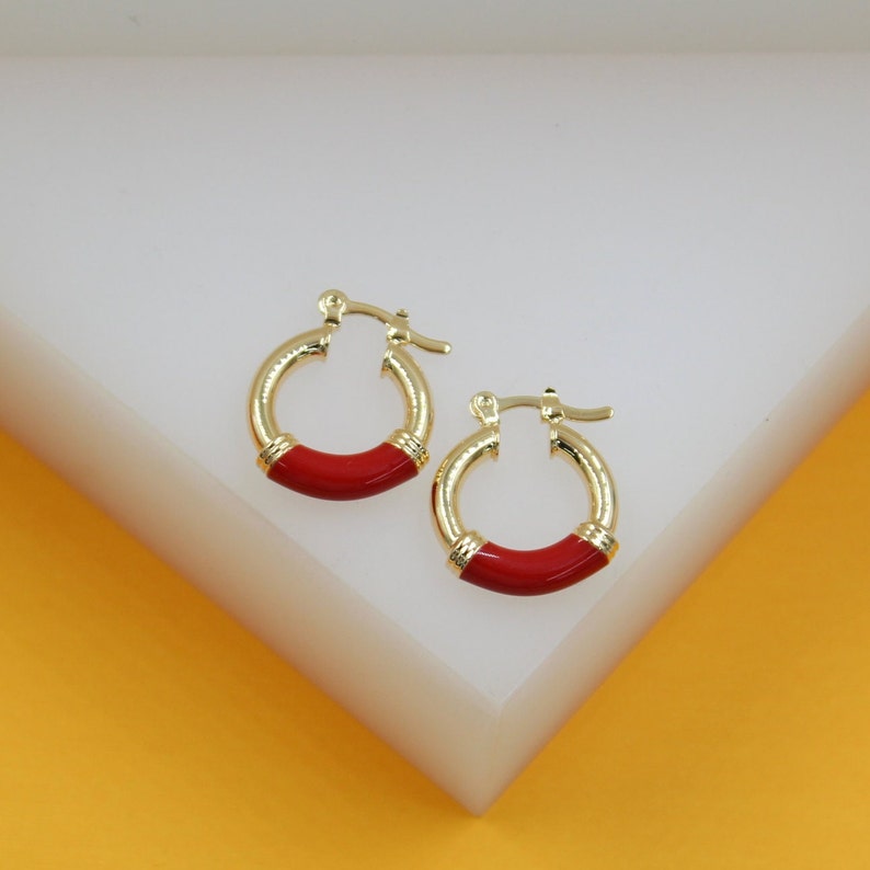 A pair of gold huggie hoop earrings featuring vibrant red enamel detailing rests on a white display surface.