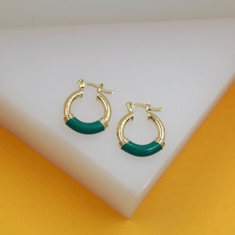 A pair of gold hoop earrings featuring vibrant green enamel accents rests on a white display stand.
