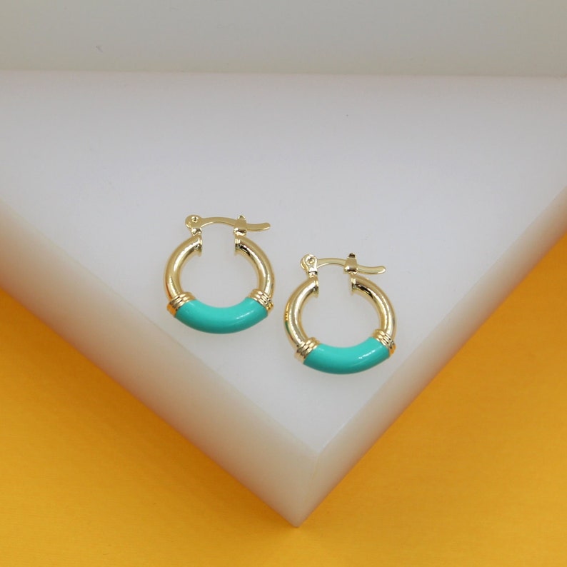 A pair of gold huggie hoop earrings featuring vibrant turquoise enamel details rests neatly on a white display surface.