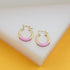 A pair of gold hoop earrings featuring pink enamel accents rest on a white display surface against a yellow background.