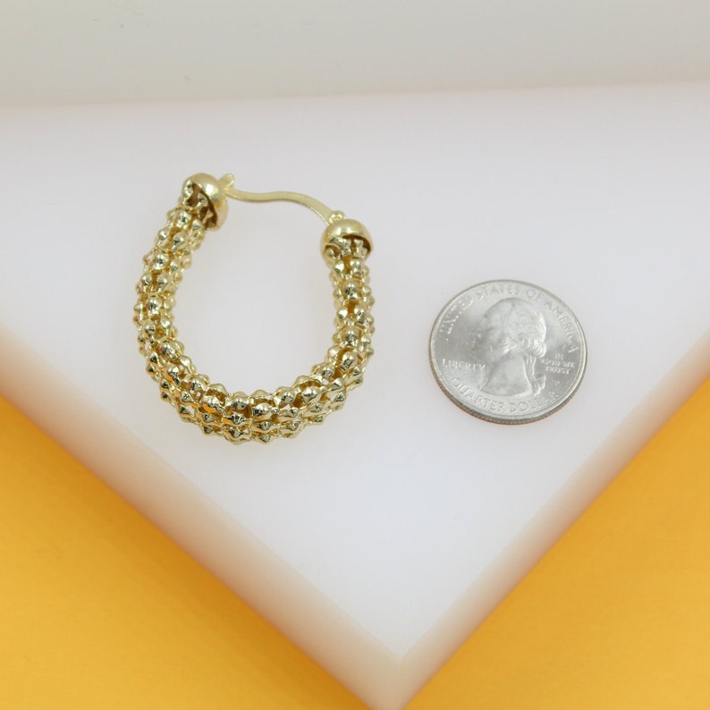 A textured gold hoop earring rests beside a US quarter for size comparison on a white surface.