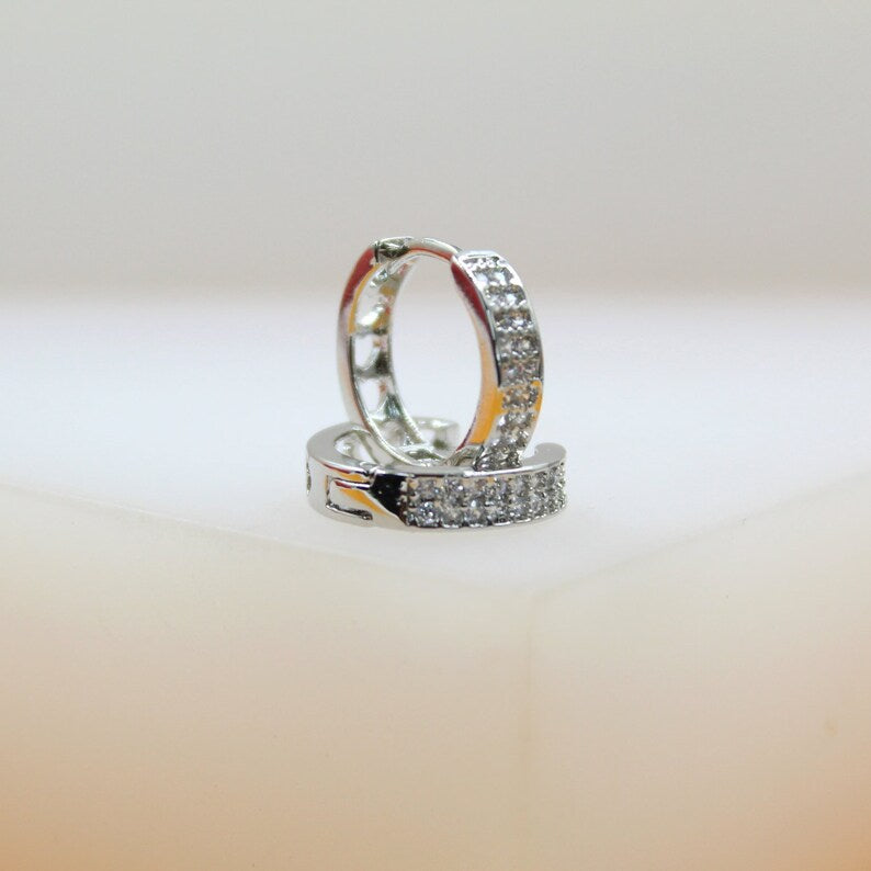A pair of sparkling silver huggie hoop earrings encrusted with small clear crystals rests on a white surface.