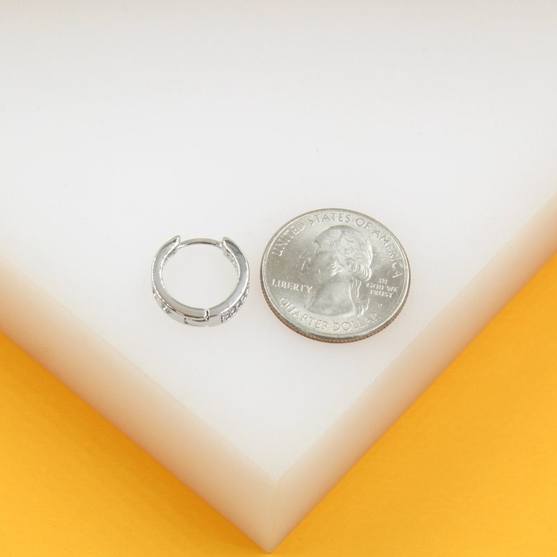 A small silver huggie hoop earring rests next to a US quarter coin on a white surface to show scale.