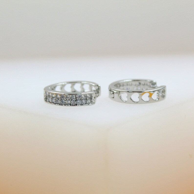 Two silver huggie hoop earrings sit on a neutral surface, showcasing a sparkling pavé crystal front on one and delicate cut-out hearts along the side of the other.