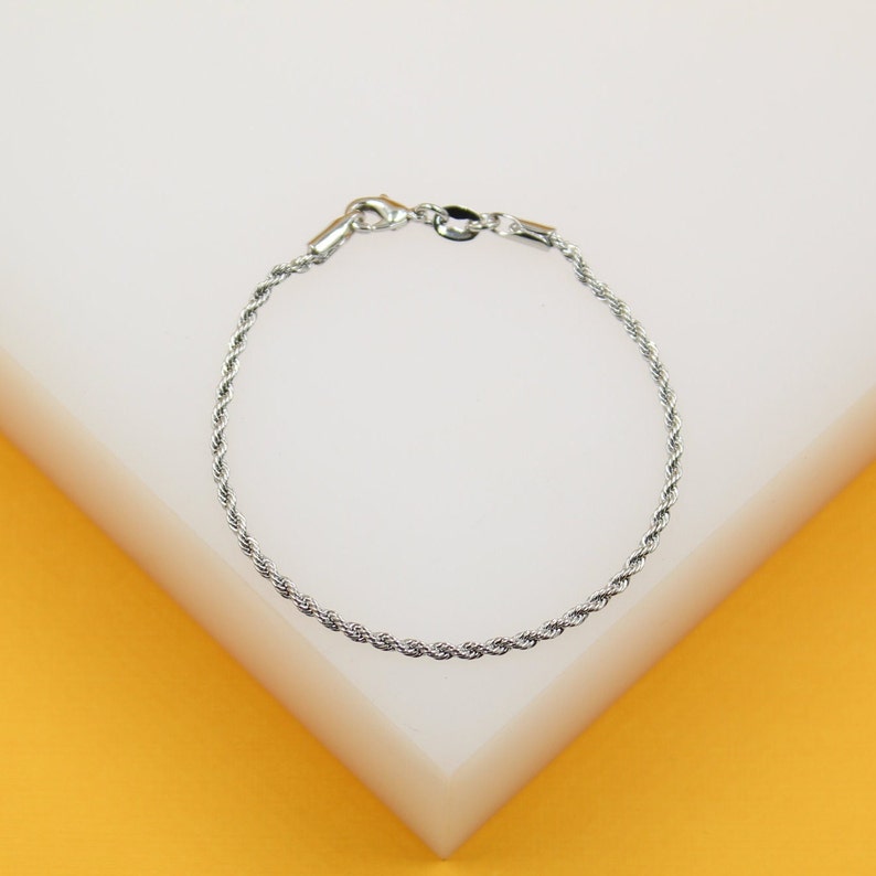A delicate silver rope chain bracelet rests on a white and yellow display stand.