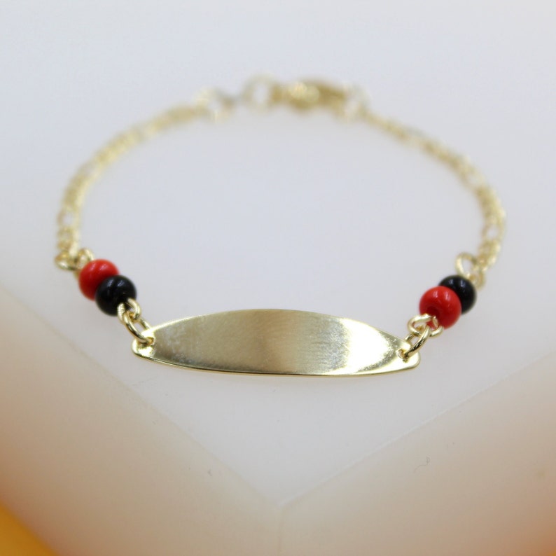 A delicate gold ID bracelet rests on a white surface, featuring a blank nameplate accented by red and black beads.