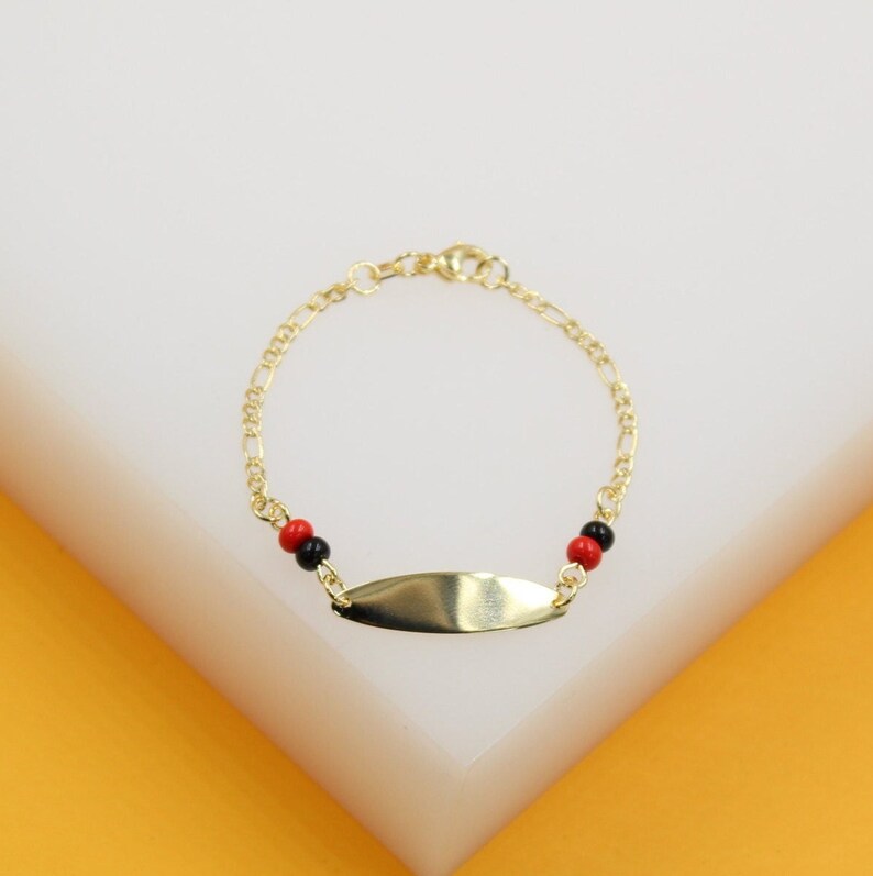 A delicate gold chain bracelet features an oval blank ID plate flanked by small red and black beads, displayed on a clean white surface.