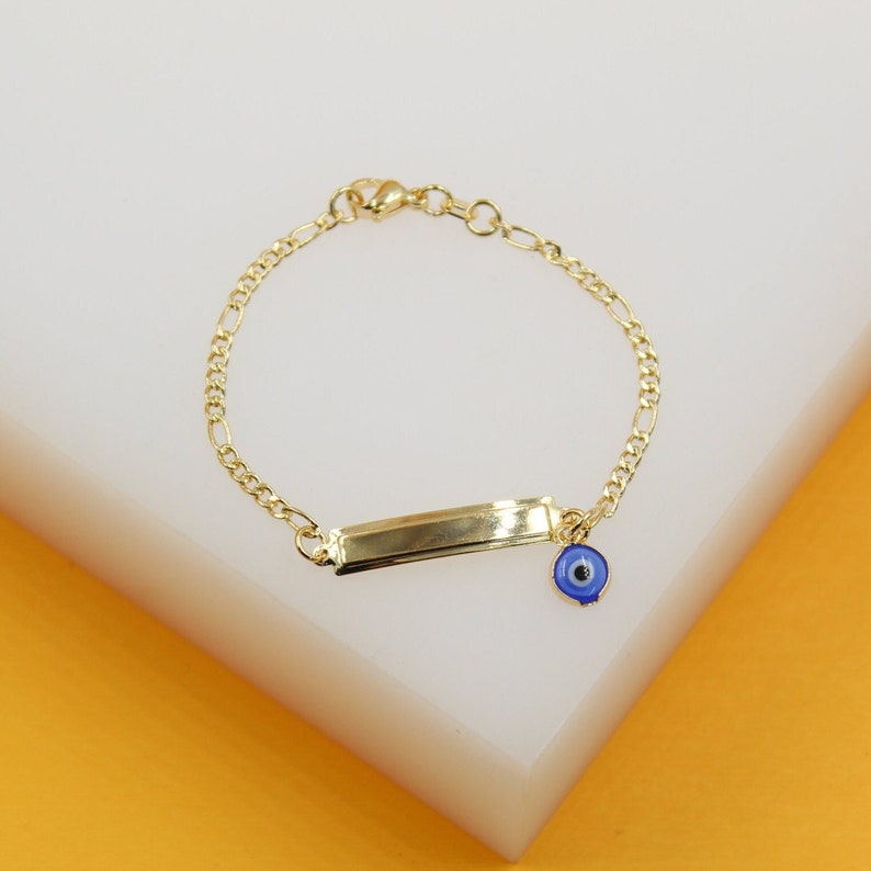 A delicate gold chain bracelet features an engraved ID bar and a dangling blue evil eye charm resting on a white display block.