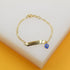 A delicate gold chain bracelet features an engraved ID bar and a dangling blue evil eye charm resting on a white display block.