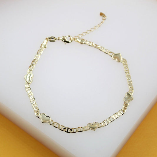A delicate gold Mariner chain anklet featuring alternating heart charms lies on a neutral white and yellow surface.