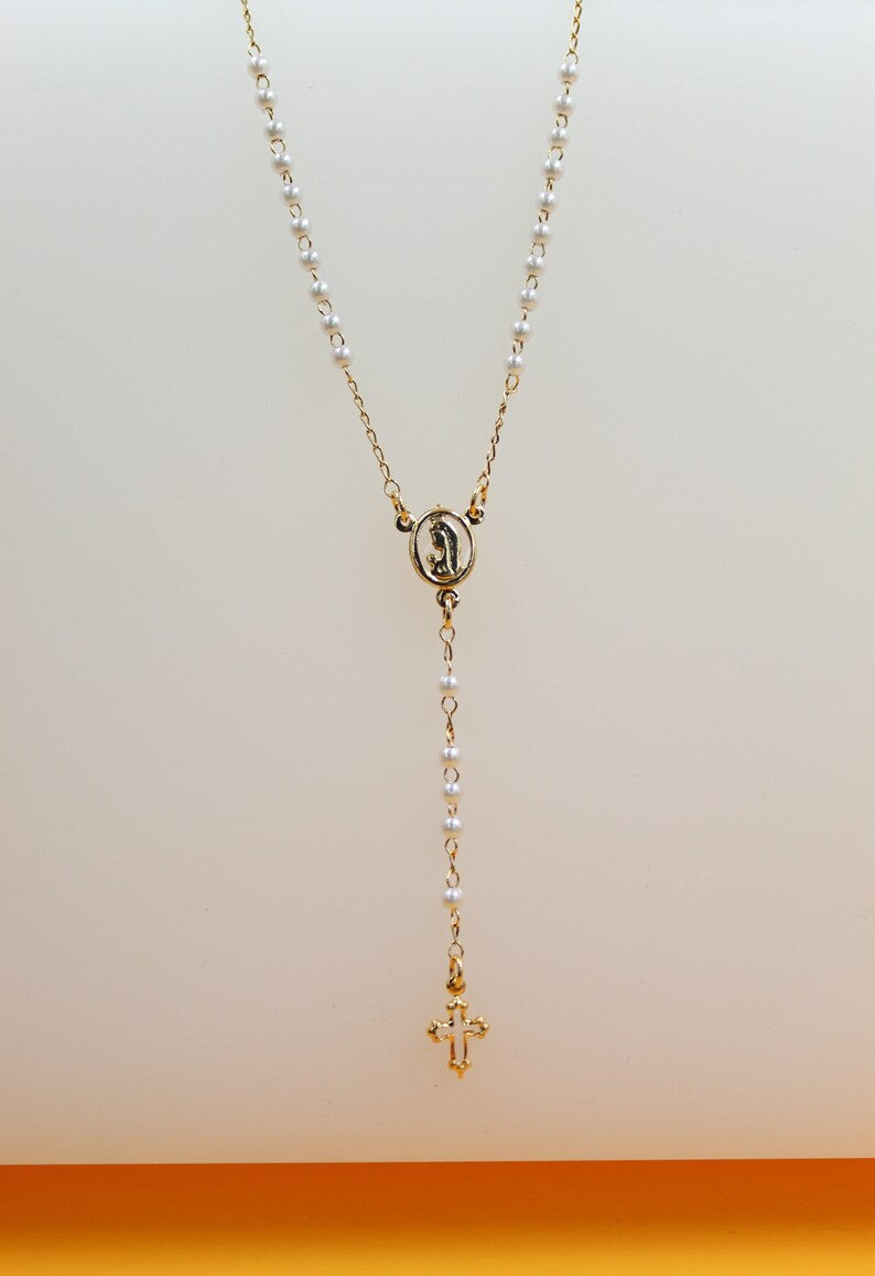 A delicate gold rosary necklace featuring white pearls and a matching crucifix rests against a neutral background.