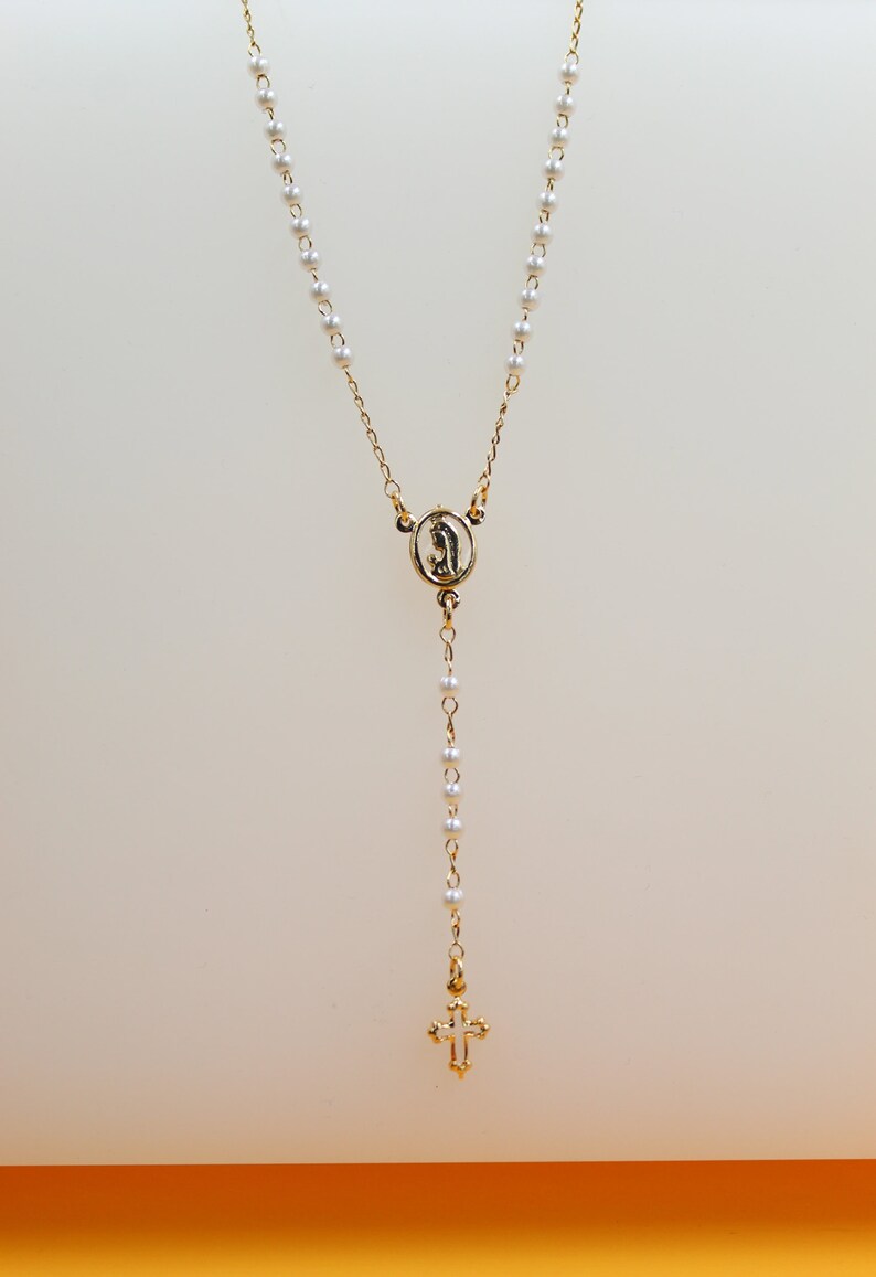 A delicate gold rosary necklace featuring white pearl beads, a central centerpiece, and a small crucifix hangs against a neutral background.