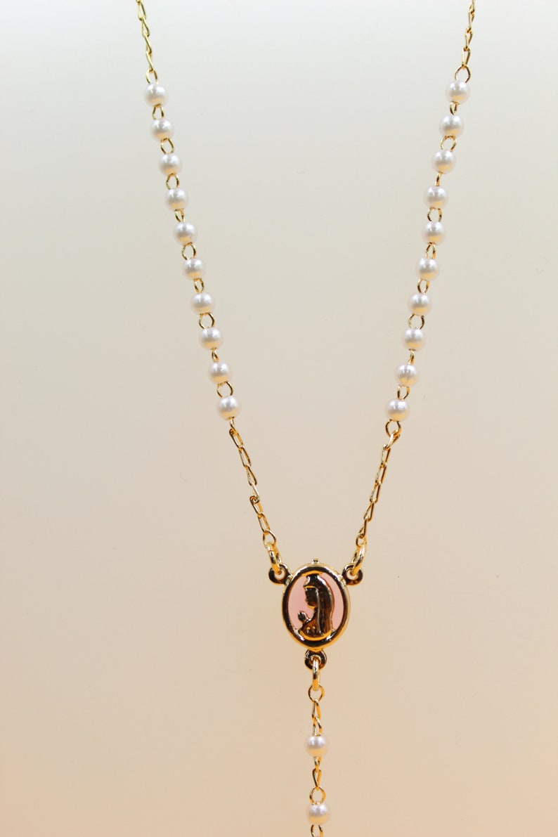 A delicate gold rosary necklace features small white pearls and an oval pink pendant depicting the Virgin Mary.