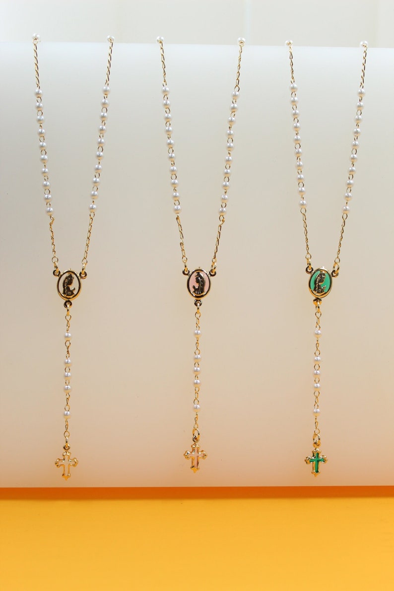 Three gold pearl rosary necklaces with colorful religious pendants and matching crosses hang vertically on a light background.